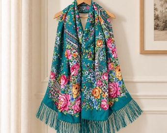 Puede incluir: Una bufanda rectangular de color verde azulado con un vibrante estampado floral de grandes rosas rosas, naranjas y amarillas, flores azules más pequeñas y hojas verdes. La bufanda termina con largos flecos a juego en el borde.