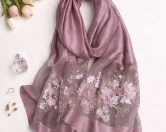 Puede incluir: Una bufanda de color malva con detalles florales bordados. La bufanda tiene una sección transparente bordada con flores blancas y rosas. La bufanda está drapeada, mostrando su textura y diseño. Un frasco de perfume y joyas están en el fondo.