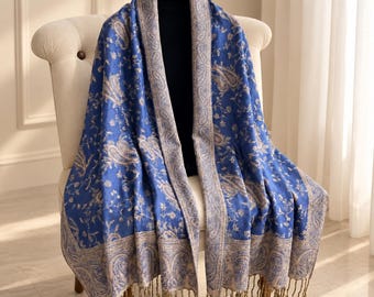 Puede incluir: Un chal azul con un patrón de cachemira y flores en color beige, que incluye un borde decorativo ancho. La tela tiene una textura suave y está rematada con flecos largos y finos de color beige en los bordes inferiores, sobre una prenda oscura.