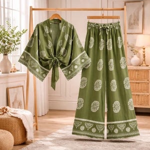 May include: Olive green two-piece outfit with a kimono-style top and wide-leg pants. The set features a white floral and geometric pattern. The top has a tie closure, and the pants have a drawstring waist. The outfit is displayed on a wooden clothing rack.