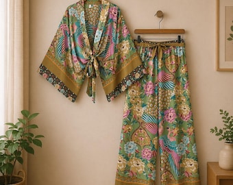 Könnte beinhalten: Ein zweiteiliges Set aus grünem Kimono-Oberteil und Hose mit weitem Bein und floralem Muster. Das Set zeigt ein lebendiges Muster aus rosa, blauen und gelben Blumen und Pfauenfedern. Der Kimono hat einen Bindverschluss und die Hose einen Kordelzug in der Taille. Das Set wird auf einem Holzbügel präsentiert.