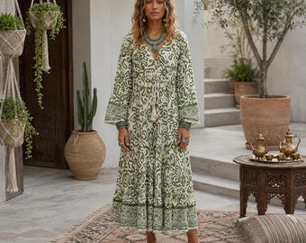 Puede incluir: Un vestido de manga larga hasta los tobillos con una base color crema y un intrincado estampado de cachemira y flores en verde oliva. La prenda tiene un escote en V, una cintura con cordón y borlas, y una falda escalonada con un dobladillo ancho.