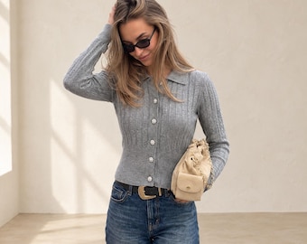 Puede incluir: Un cárdigan gris de manga larga con textura acanalada, cuello y cinco botones circulares con incrustaciones de cristal, combinado con vaqueros azules, un cinturón de cuero negro con hebilla dorada y un bolso beige con cordón y cadena dorada.
