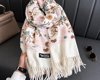 Puede incluir: Una bufanda de color crema con un intrincado patrón floral en tonos rosa, marrón y azul. La bufanda tiene una etiqueta rectangular negra con un icono de cabra y el texto Cashmere 100% Wool, terminada con largos flecos en los extremos.