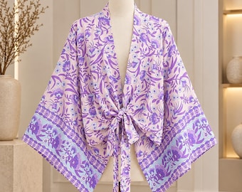 Puede incluir: Un kimono rosa claro con un vibrante estampado floral en tonos morados y azules con motivos botánicos de hojas. La prenda tiene mangas anchas, un diseño frontal abierto y una lazada de tela en la cintura para un cierre anudado en el centro.