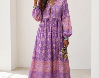 Puede incluir: Un vestido midi de manga larga en tonos morados y rosa claro, con un estampado floral repetitivo en toda la prenda. El vestido tiene un escote en V con finos lazos, una cintura fruncida y mangas sueltas con puños elásticos en las muñecas.