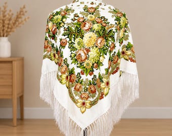 Puede incluir: Un chal cuadrado de color blanco con un estampado floral vibrante en tonos naranja, amarillo y verde. La tela presenta ilustraciones detalladas de rosas y otras flores, rematado con largos flecos blancos en los bordes.