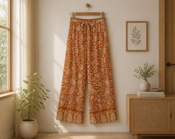 Puede incluir: Pantalones de pierna ancha en color naranja quemado con un estampado floral y de cachemira blanco en toda la prenda. Los pantalones tienen una cintura elástica con un cordón de tela a juego y un borde decorativo ancho en el dobladillo.