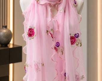 Puede incluir: Una bufanda rosa claro semitransparente con patrones florales bordados en tonos rojo, morado y verde. La bufanda tiene un borde ondulado con volantes y un remate rosa a juego, colocada sobre un maniquí para mostrar la textura de la tela.
