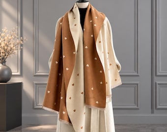 Puede incluir: Una bufanda reversible con un diseño bicolor en marrón y crema. La bufanda está decorada con un patrón repetitivo de pequeñas formas de corazón simples en toda la superficie. Tiene un dobladillo recto con flecos y está sobre un maniquí.