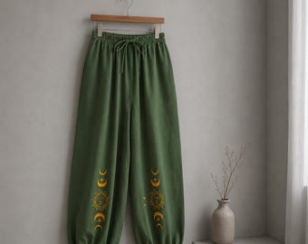 Puede incluir: Pantalones holgados de color verde oliva con cintura elástica y cordón ajustable. La parte inferior de cada pernera presenta un estampado gráfico vertical en amarillo que representa una serie de motivos celestiales de soles, lunas y estrellas.