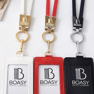 Puede incluir: Tres portatarjetas de identificación verticales con cordones a juego en blanco, rojo y negro. Cada soporte presenta el logotipo de BOASY SUPPLY COMPANY. Los cordones se unen a los soportes mediante cierres metálicos y anillas.