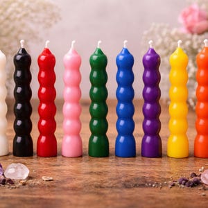 May include: A selection of eight brightly coloured pillar candles. The candles are white, black, red, pink, green, blue, purple, yellow, and orange. Each candle has a distinctive, textured design and a white wick.