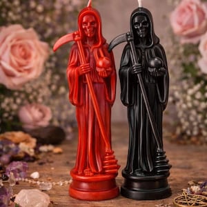 Op de afbeelding: Twee Grim Reaper-kaarsen. Eén is rood, de andere zwart. Elke figuur houdt een zeis en een bol vast. De kaarsen staan op een houten oppervlak met bloemen op de achtergrond. De kaarsen zijn ongeveer 18 cm hoog.