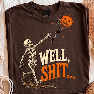 May include: A dark brown t-shirt featuring a graphic of a white skeleton reaching toward an orange jack-o'-lantern balloon. The text "WELL, SHIT..." is printed in large, bold, cream and orange letters below the skeleton on the front of the shirt.