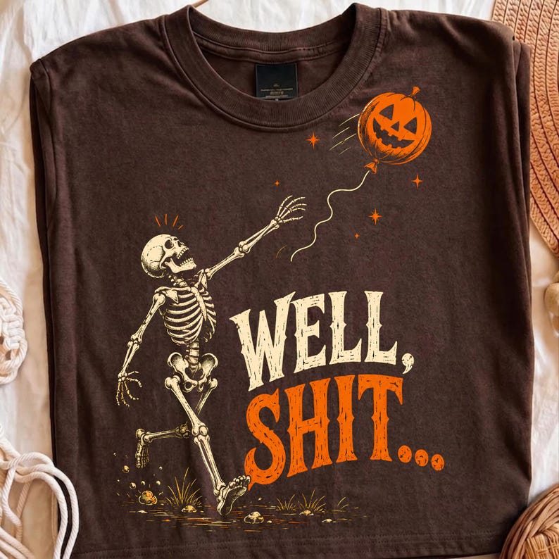 May include: A dark brown t-shirt featuring a graphic of a white skeleton reaching toward an orange jack-o'-lantern balloon. The text "WELL, SHIT..." is printed in large, bold, cream and orange letters below the skeleton on the front of the shirt.