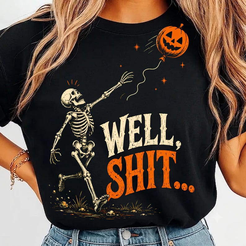 May include: A black t-shirt featuring a graphic of a skeleton reaching for a floating jack-o'-lantern balloon. The text "WELL, SHIT..." is printed in large, distressed cream and orange lettering below the skeleton, with small stars in the background.