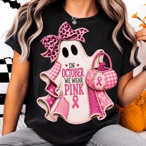 Peut inclure: Un t-shirt noir avec un motif de fantôme blanc portant un nœud à imprimé léopard rose et tenant un panier citrouille rose. Le texte indique IN OCTOBER WE WEAR PINK, avec un ruban rose. Le fantôme présente divers empiècements à motifs roses.