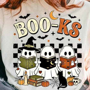 Peut inclure: Un t-shirt couleur crème avec un motif portant l'inscription "BOO-KS" au-dessus de trois fantômes blancs lisant des livres. Le design comprend un chapeau de sorcière noir, un croissant de lune, des chauves-souris, des étoiles, des livres, une bougie, des citrouilles, un chat noir et une tasse en forme de crâne.