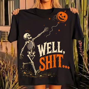 May include: A black t-shirt featuring a graphic of a skeleton reaching for a floating orange jack-o'-lantern balloon. The text "WELL, SHIT..." is printed in large, stylized white and orange lettering below the skeleton on the front of the shirt.