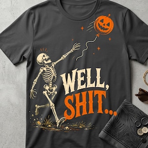 May include: A dark gray t-shirt featuring a graphic of a skeleton reaching for a floating jack-o'-lantern balloon. The text "WELL, SHIT..." is printed in large, stylized cream and orange letters below the skeleton, with small orange stars.