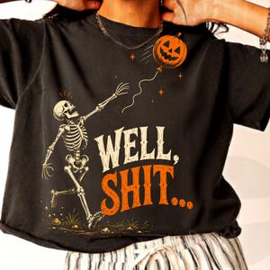 May include: A black short-sleeve t-shirt featuring a graphic of a skeleton reaching for a floating jack-o'-lantern balloon. The text "WELL, SHIT..." is printed in white and orange lettering below the skeleton. The design includes small orange stars.