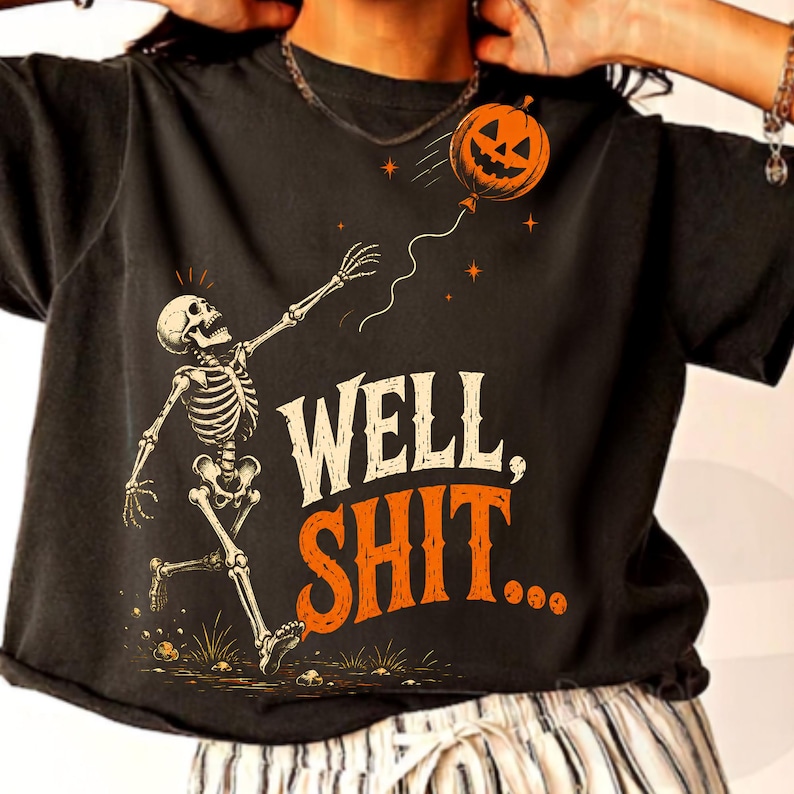 May include: A black short-sleeve t-shirt featuring a graphic of a skeleton reaching for a floating jack-o'-lantern balloon. The text "WELL, SHIT..." is printed in white and orange lettering below the skeleton. The design includes small orange stars.