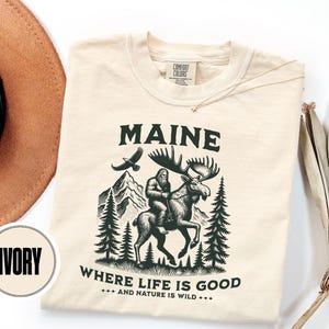 May include: Ivory-colored t-shirt with a graphic of a Sasquatch riding a moose, mountains, and pine trees. The text "MAINE" and "WHERE LIFE IS GOOD" are printed on the shirt. A brown hat is visible in the upper left corner.