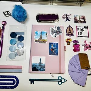 May include: A collection of stationery items, including a pink notebook with Eiffel Tower stickers, pens, a blue pom-pom keychain, and decorative paper clips. The items are arranged on a white surface.