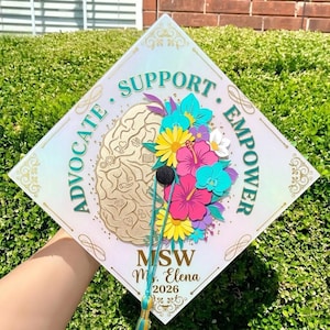 Puede incluir: Un gorro de graduación decorado con las palabras "Advocate, Support, Empower" que rodean un gráfico de cerebro y flores de papel de colores. El gorro también incluye el texto "MSW M. Elena 2026".