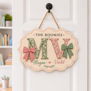 May include: A scalloped wooden wall sign featuring the text THE ROOMIES and the names Megan and Violet. The sign displays large letters M and V with floral patterns, accented by a pink bow on the left and a sage green bow on the right.