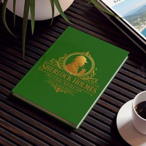 May include: A green hardcover journal featuring a gold foil silhouette of Sherlock Holmes and the text Sherlock Holmes Case Notes 221B Baker Street. The journal is placed on a dark slatted table next to a white coffee cup and a travel magazine.