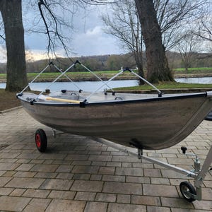 Può includere: Una barca in alluminio argentato su un rimorchio con una ruota rossa. La barca ha un telaio bianco e un remo di legno. La barca è parcheggiata su una superficie di mattoni vicino a un fiume.