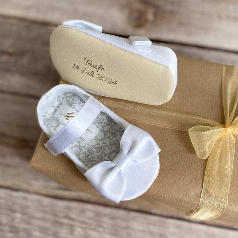 May include: White satin baby shoes with a bow on the front. The sole of the shoe is cream colored and has the text "Taufe 14 Juli 2024" written on it.