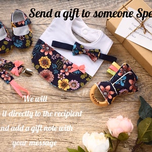 May include: A gift set for a baby, featuring a floral print bib, a bow tie, a pair of shoes, a teether, and a pacifier clip. The set is packaged in a brown box with a gift note.