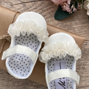 K&ouml;nnte beinhalten: Ein Paar wei&szlig;e Satin-Babyschuhe mit wei&szlig;er Spitzenborte und grauer Tupfen-Innenausstattung. Die Schuhe liegen auf einem braunen Papieruntergrund.