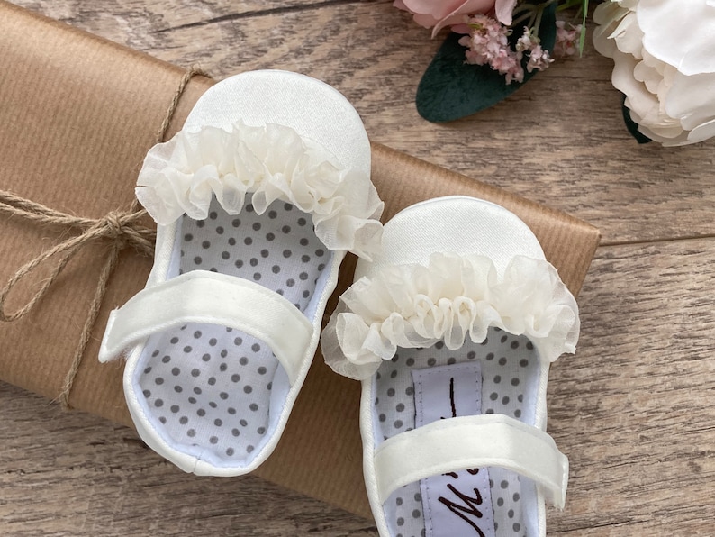 K&ouml;nnte beinhalten: Ein Paar wei&szlig;e Satin-Babyschuhe mit wei&szlig;er Spitzenborte und grauer Tupfen-Innenausstattung. Die Schuhe liegen auf einem braunen Papieruntergrund.