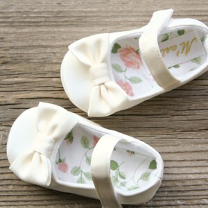 May include: A pair of white satin baby shoes with a bow on the front and a floral print lining. The shoes have a strap with a gold label that says "M. & Co."