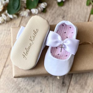 May include: White baby shoes with a satin bow and a small silver cross charm. The shoes are personalized with the name "Helene Kathe" on the sole.
