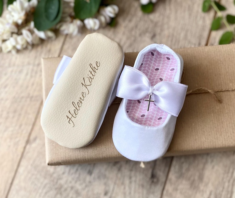 May include: White baby shoes with a satin bow and a small silver cross charm. The shoes are personalized with the name "Helene Kathe" on the sole.