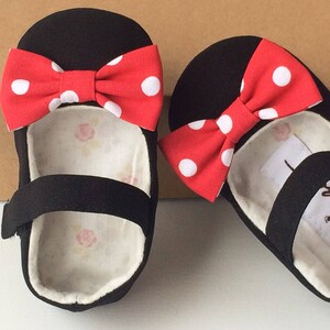 May include: A pair of black canvas baby shoes with red and white polka dot bows. The shoes have a strap across the top and a white lining.