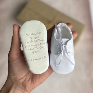 Puede incluir: Un par de zapatos blancos para bebé. La suela de un zapato tiene el texto "For this child I prayed and the Lord hath given me my petition which I asked of him" y "1 Samuel 1.27". El otro zapato tiene un lazo de cinta.
