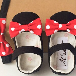 May include: Black baby shoes with red and white polka dot bows. The shoes have a black strap across the top and a white label with the text "M'an" on the inside. A matching red and white polka dot bow tie is included.