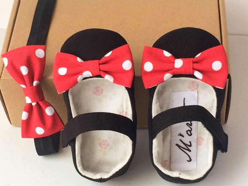 May include: Black baby shoes with red and white polka dot bows. The shoes have a black strap across the top and a white label with the text "M'an" on the inside. A matching red and white polka dot bow tie is included.