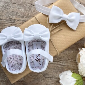 May include: White baby shoes with a bow on the front and a white headband with a bow. The shoes have a white satin bow and the headband has a white satin bow. The shoes are on a brown box with a white label that says "Mr."