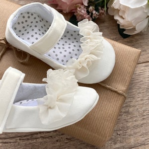 K&ouml;nnte beinhalten: Zwei wei&szlig;e Babyschuhe mit einer wei&szlig;en Spitzenr&uuml;sche vorne. Die Schuhe liegen auf einem braunen Papierpaket.