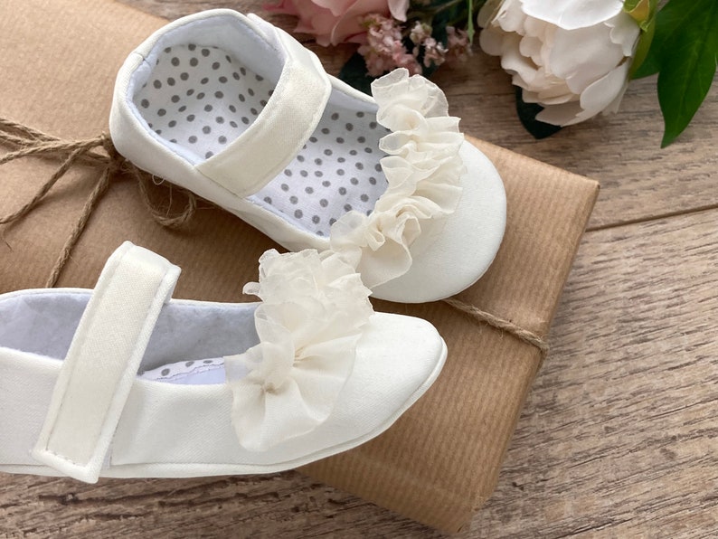 K&ouml;nnte beinhalten: Zwei wei&szlig;e Babyschuhe mit einer wei&szlig;en Spitzenr&uuml;sche vorne. Die Schuhe liegen auf einem braunen Papierpaket.