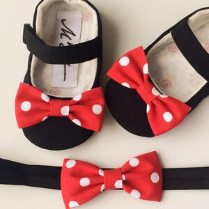 May include: A set of black baby shoes with red and white polka dot bows. The shoes are paired with a black headband with a matching red and white polka dot bow.