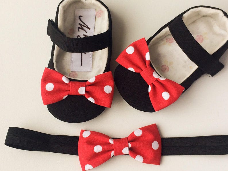 May include: A set of black baby shoes with red and white polka dot bows. The shoes are paired with a black headband with a matching red and white polka dot bow.