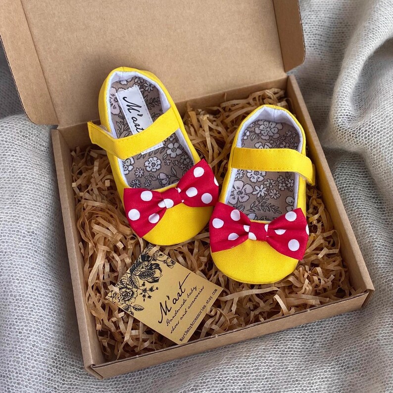 K&ouml;nnte beinhalten: Ein Paar gelbe Babyschuhe mit roten und wei&szlig;en Tupfen-Schleifen. Die Schuhe befinden sich in einer braunen Schachtel mit zerkn&uuml;lltem Papier. Die Schachtel hat ein Etikett mit der Aufschrift "M'art Handgemachte Babyschuhe und Accessoires"